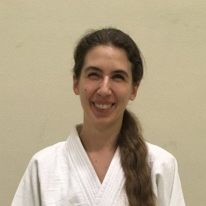 Sally, Eine Frau mit langen, zu einem Pferdeschwanz gebundenen braunen Haaren, lächelt herzlich. Sie trägt einen weißen Kampfkunst-Gi. Ihr Gesichtsausdruck ist fröhlich und entspannt, mit leicht zusammengekniffenen Augen in freundlicher Weise. Der Hintergrund ist eine schlichte, hellfarbige Wand.