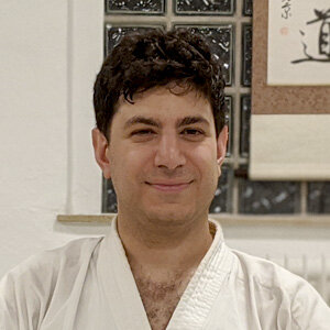 Ein Mann mit kurzen, dunklen lockigen Haaren lächelt sanft. Er trägt einen weißen Karate-Gi, eine traditionelle Uniform. Hinter ihm befindet sich eine Wand mit Glasbausteinen und eine Schriftrolle mit asiatischen Schriftzeichen. Die Umgebung scheint drinnen zu sein, möglicherweise ein Dojo oder ein Trainingsbereich für Kampfsport.