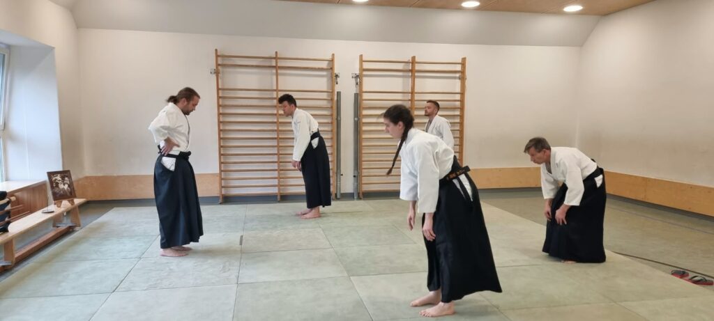 Fünf Aikidoka in weißen Jacken und schwarzen Hakama-Hosen wärmen sich gemeinsam auf einer gepolsterten Dojo-Fläche auf. Alle sind barfuß und beugen sich nach vorne, als würden sie Rücken und Beine dehnen. Die Gruppe besteht aus Männern und einer Frau mit einem Altersbereich von jungen Erwachsenen bis mittlerem Alter: Eine jüngere Frau mit langem dunklem Zopf im Vordergrund, ein jüngerer Mann mit kurzen dunklen Haaren in der Mitte, zwei mittelalte Männer mit heller Haut und kurzen Haaren rechts und im Hintergrund sowie ein weiterer mittelalter Mann mit heller Haut und zu einem Zopf gebundenen langen Haaren links. Ihre Körperformen reichen von schlank bis durchschnittlich, und sie stehen locker über die Matte verteilt statt in einer starren Reihe, was dem Aufwärmen eine entspannte, informelle Stimmung verleiht. Hinter ihnen befinden sich Sprossenwände und schlichte weiße Wände. Auf einer niedrigen Bank links steht ein gerahmtes Schwarzweiß-Porträt von O Sensei, dem Begründer des Aikido, das die traditionelle Atmosphäre des Dojos unterstreicht.