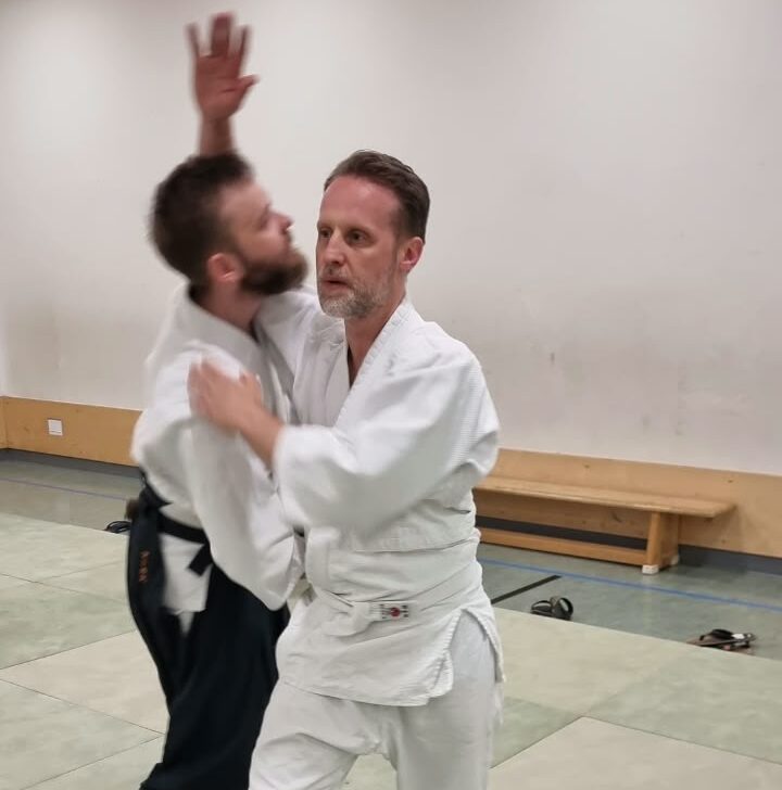Zwei Aikido-Schüler üben die Technik Irimi Nage auf Matten im Dojo. Vorn steht ein hellhäutiger Anfänger in einem weißen Gi mit weißer Hose und weißem Gürtel, barfuß in einer stabilen Stellung mit dem linken Bein vorn und dem rechten Bein hinten, die Knie leicht gebeugt. Sein Oberkörper ist leicht nach rechts gedreht, während er tief eintritt und seinen Partner aus dem Gleichgewicht bringt. Sein rechter Arm liegt um den oberen Rücken und Schulterbereich des fortgeschrittenen Schülers, sein linker Arm kontrolliert tiefer in Höhe der Rippen oder Taille. Sein Gesichtsausdruck ist konzentriert und ruhig, sein Blick geht leicht nach vorn links statt direkt zu seinem Partner. Der fortgeschrittene Schüler, ebenfalls hellhäutig, trägt ein weißes Gi-Oberteil und eine dunkle Hakama-Hose, was sein höheres Niveau zeigt. Er wird nach links aus dem Gleichgewicht gebracht, sein Oberkörper lehnt sich nach vorn in die Brust des Anfängers. Sein linker Arm ist gebeugt und hoch in der Nähe seines Kopfes mit geöffneter Hand, als wäre ein Schlag umgelenkt worden, während sein rechter Arm unten und größtenteils nicht sichtbar ist. Beide stehen barfuß auf großen, quadratischen Matten in hellem Grün und Beige. Im Hintergrund befindet sich ein schlichtes, helles Dojo mit einer glatten Wand, einer niedrigen Holzbank entlang der Wand, einigen Gegenständen wie Gürteln auf dem Boden und hellem Licht von Deckenlampen.
