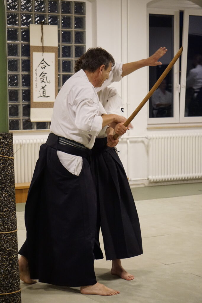 Zwei Personen üben Kampfsport in einem Dojo. Sie tragen traditionelle Kampfsportkleidung mit weißen Oberteilen und schwarzen Hakama Hosen. Die Person rechts hält ein Holzschwert, bekannt als Bokken, während die Person links sie anscheinend anleitet oder unterrichtet. Im Hintergrund ist eine Ausstellung mit mehreren Holzschwertern und eine Schriftrolle mit japanischen Schriftzeichen an der Wand zu sehen. Der Raum hat ein sauberes, minimalistisches Design mit weißen Wänden und einem Spiegel, der einen Teil der Szene reflektiert.